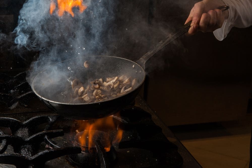 Home Image: Side view mushroom frying with smoke and fire and human hand and pan in stove