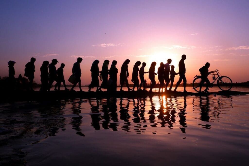 Home Image: A line of silhouetted people walking along a waterfront at sunset with reflections on the water