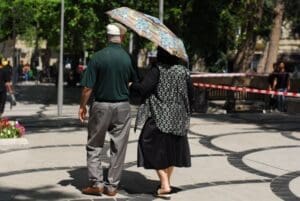 Home Image: An elderly couple strolls through a sunny park, shading themselves with an umbrella