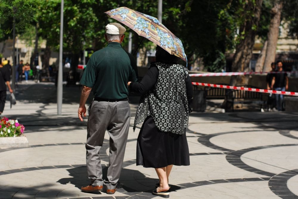 Home Image: An elderly couple strolls through a sunny park, shading themselves with an umbrella