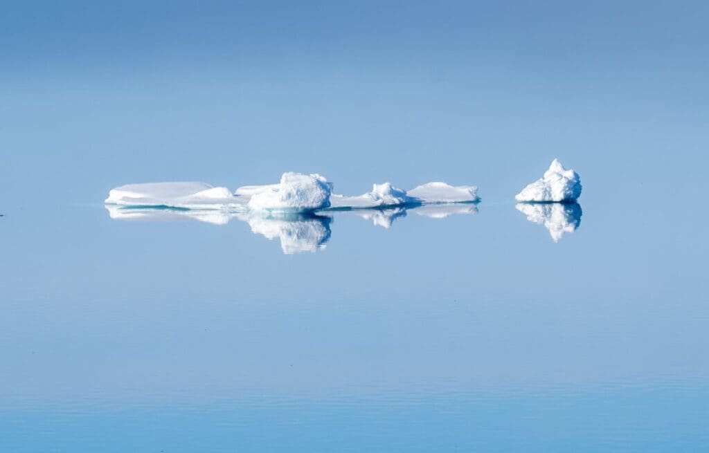 Home Image: Icebergs floating on calm water, with their reflections visible beneath a clear blue sky (s. climate change, climate fingerprinting)